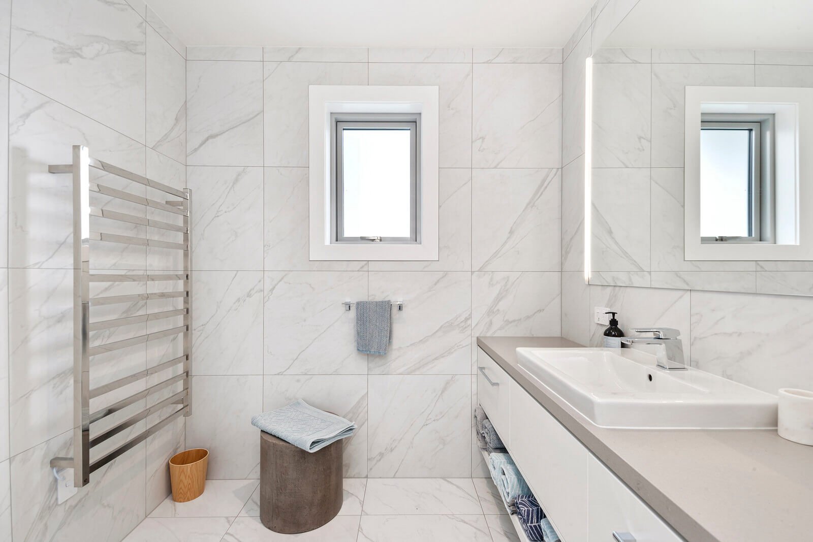 Bright modern bathroom with white marble tiles, a vanity with a sink, two windows, a towel on a towel rack, a stool with a towel, and a heated towel rail.