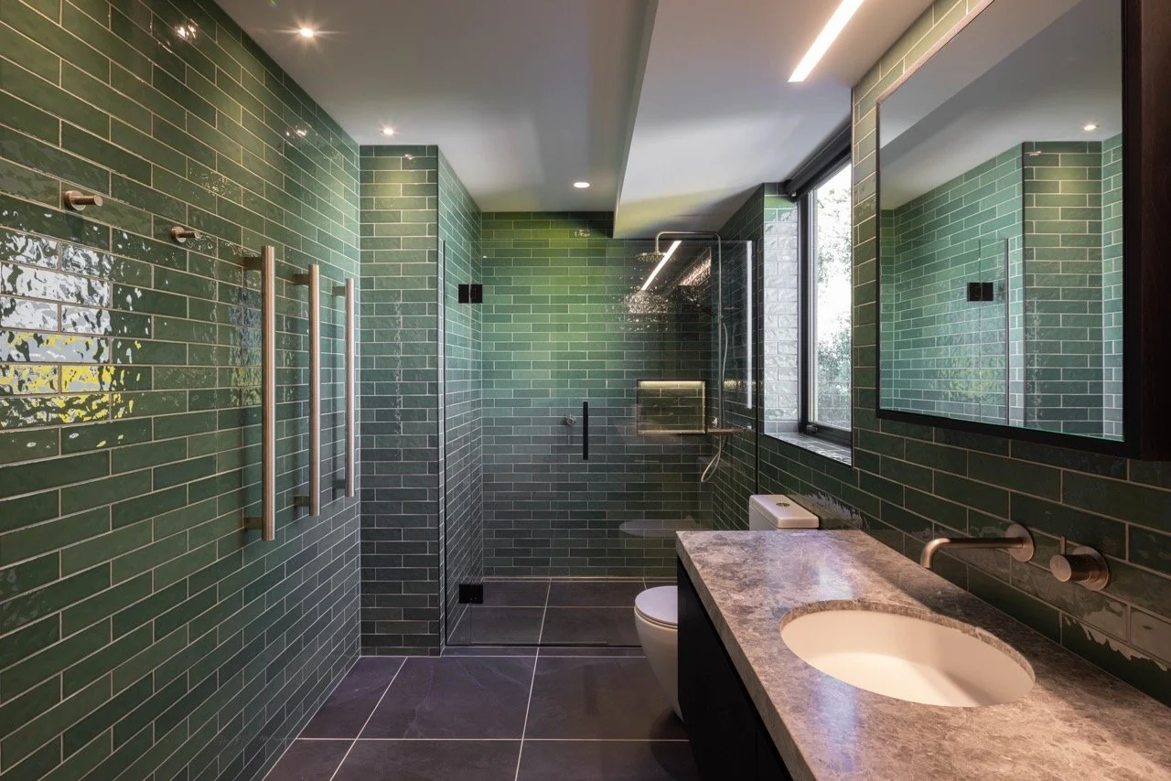 Modern bathroom with green subway tile walls, a glass-enclosed walk-in shower, a marble countertop sink, a large mirror, and a window showing outdoor greenery.