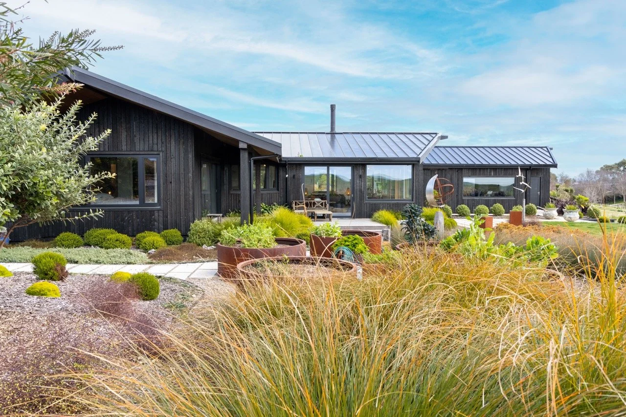 Modern black house with large windows, surrounded by a landscaped garden with various plants and grasses