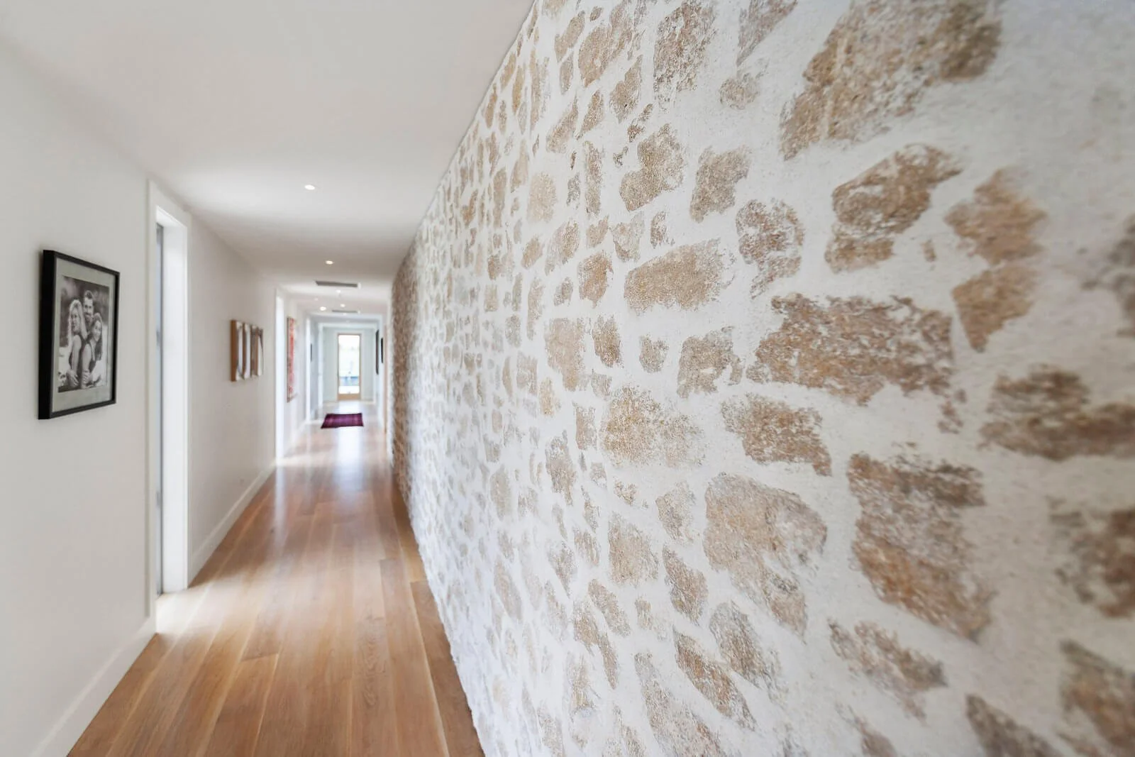Interior hallway with a textured stone wall on one side, white walls with framed photographs or artwork, wooden flooring, and a door at the end of the corridor with a window and a small red rug.