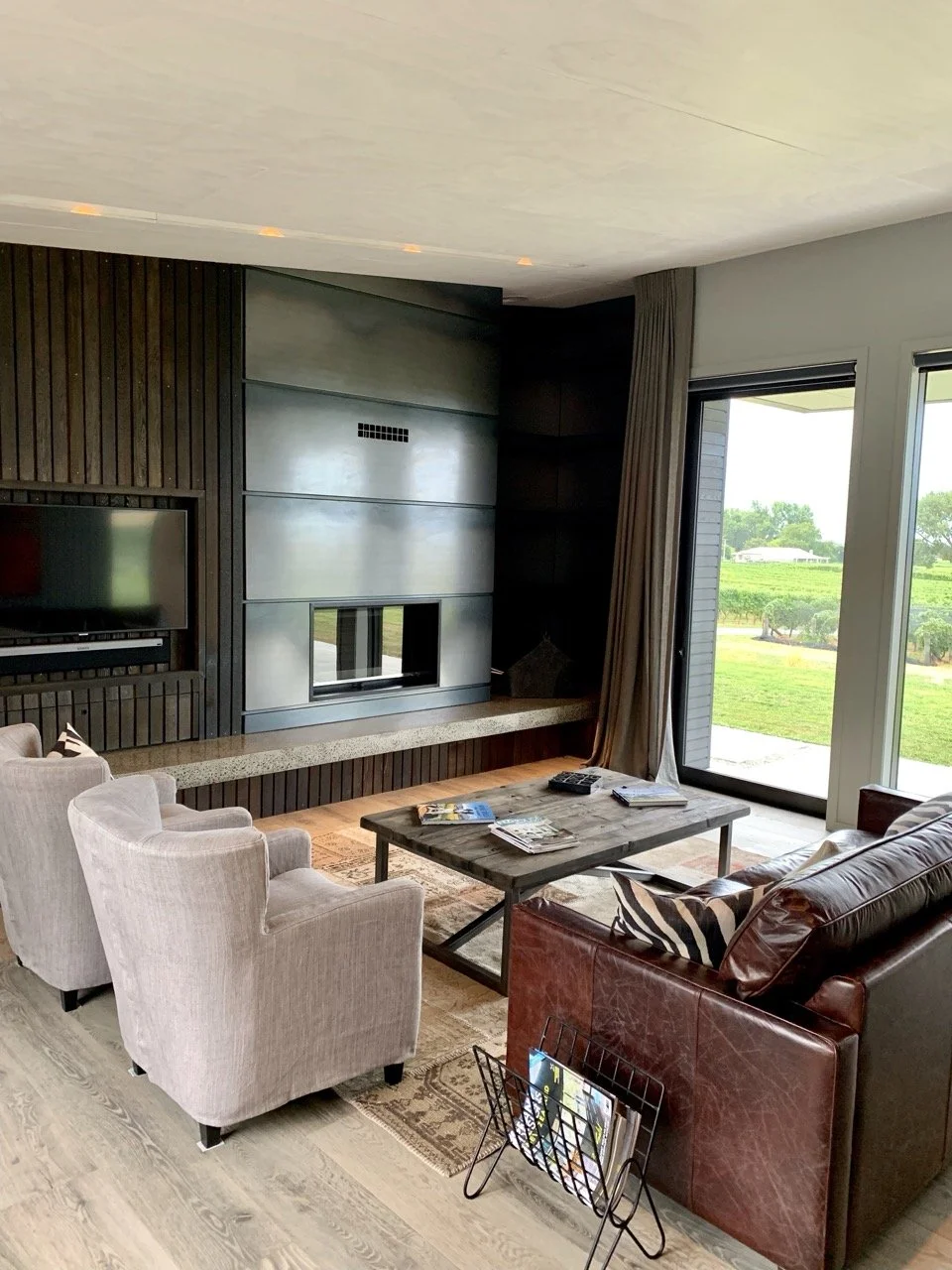 Living room with beige armchairs, a red leather sofa with pillows, a rectangular coffee table with magazines, and a flatscreen TV mounted on a dark wood paneled wall. Large glass sliding doors reveal a green outdoor landscape.