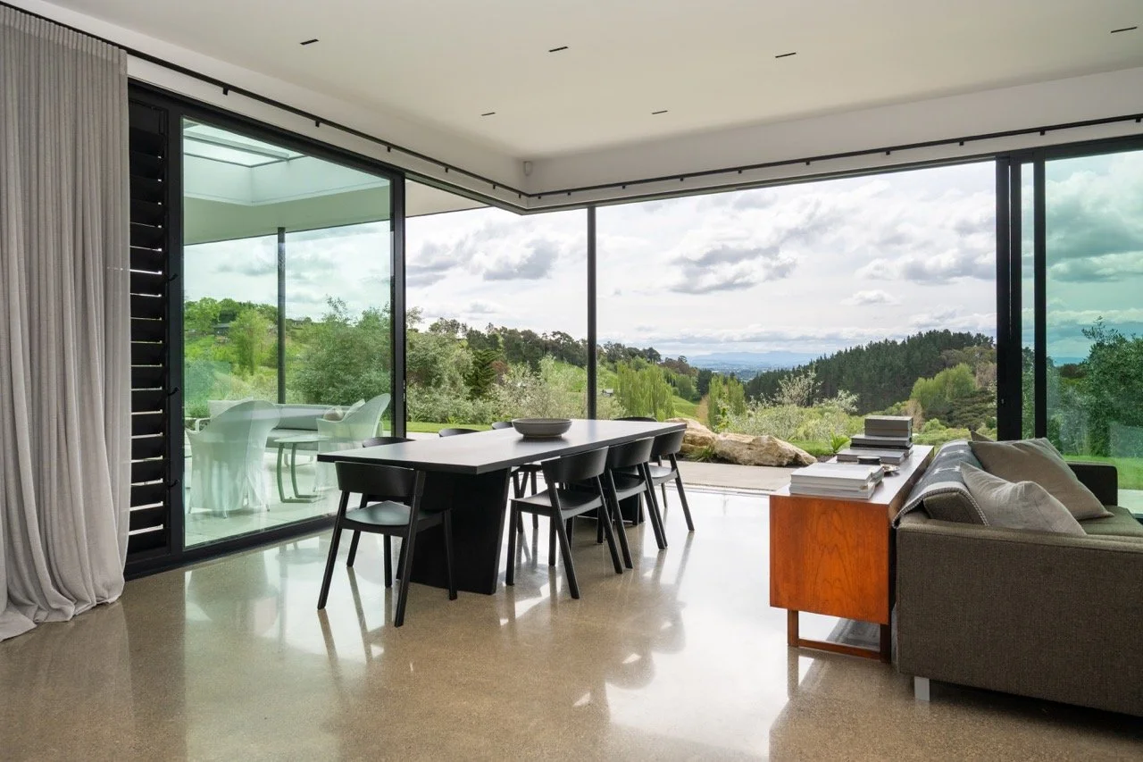 Modern living room with large floor-to-ceiling windows overlooking a lush green landscape, featuring a dining table with six black chairs, a gray sofa, a wooden side table with stacks of books, and a glass door leading to an outdoor patio.