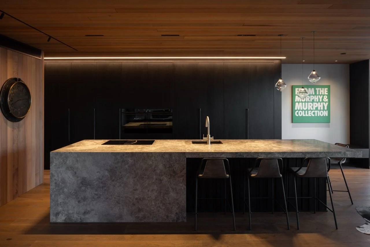 Modern kitchen with a large marble island, black cabinets, and wooden ceiling and wall accents. Three barstools are at the island, pendant lights hang above, and a green sign reading 'FROM THE MURPHY & MURPHY COLLECTION' is on the wall.