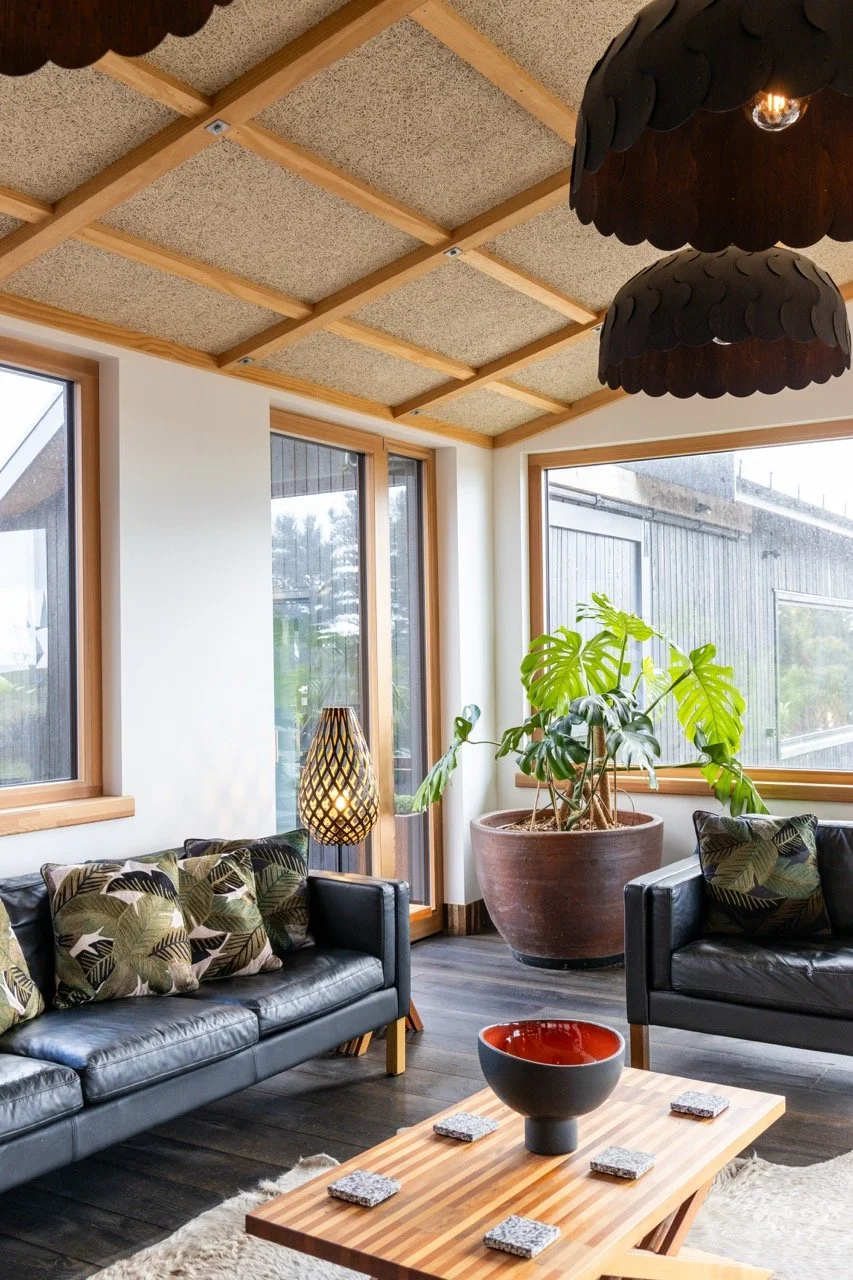 A cozy living room with large windows, black leather couches with patterned pillows, a wooden coffee table with coasters and a decorative bowl, a large potted plant, a woven floor lamp, and wooden beam ceiling panels.