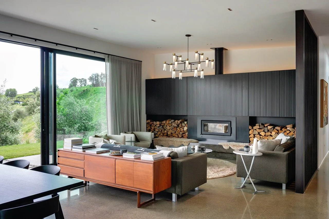 Modern living room with large window view of green landscape, black wood-paneled wall with built-in fireplace and stacked firewood, contemporary chandelier, sofas, and side tables.