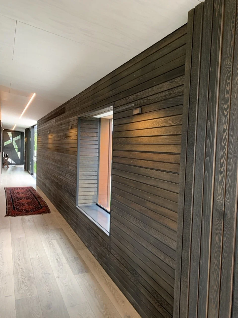 Interior hallway with wooden wall paneling, large window, light wood flooring, and a small red patterned rug.