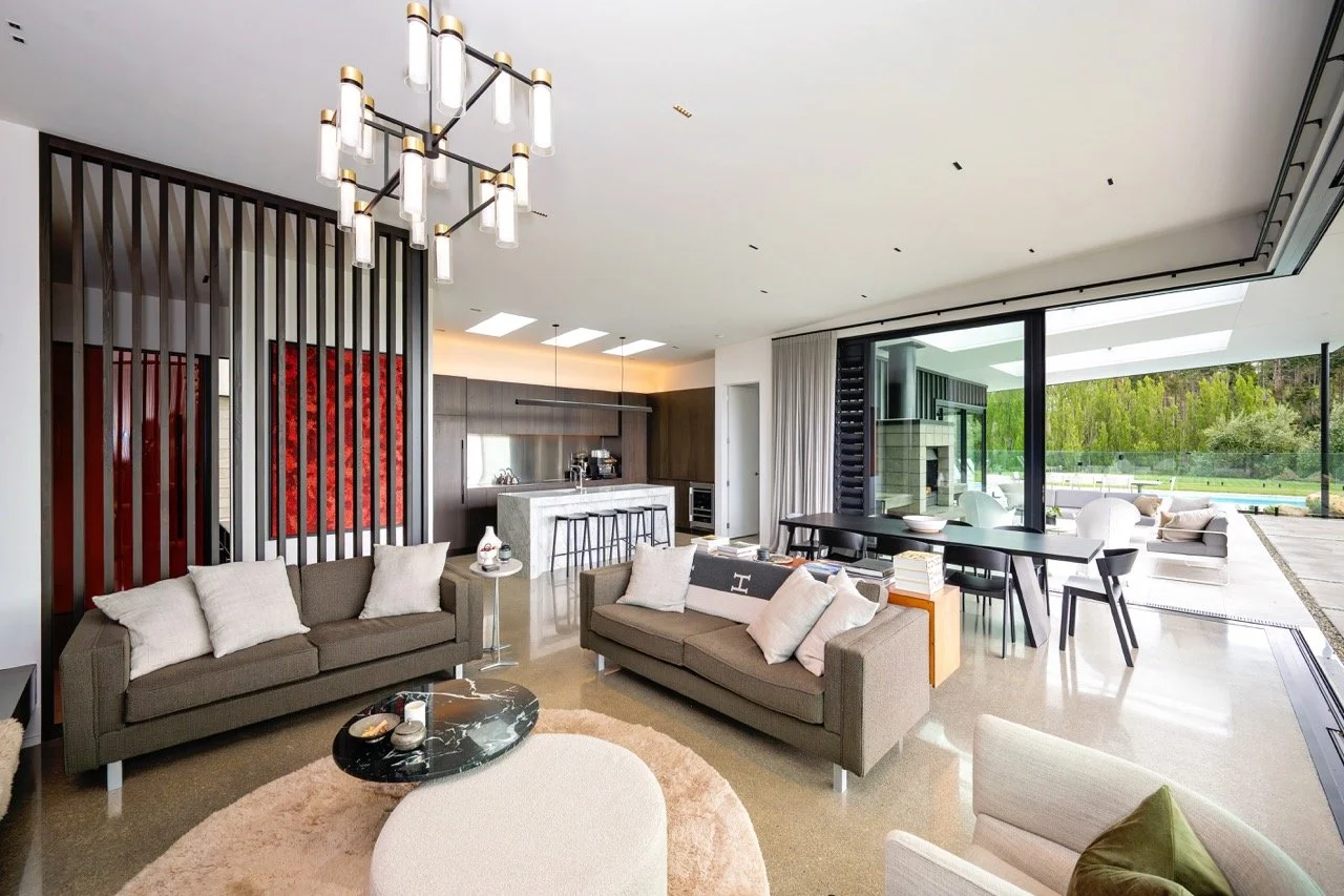 Modern open-concept living space with sofas, a marble kitchen island, dining table, and large glass sliding doors leading to an outdoor patio with view of greenery.