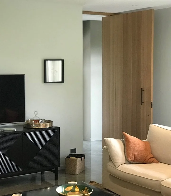 Living room with a beige sofa with brown pillows, a black TV stand with a TV, a tray with glassware, a small black and gold bowl on a table, and a wooden sliding door.
