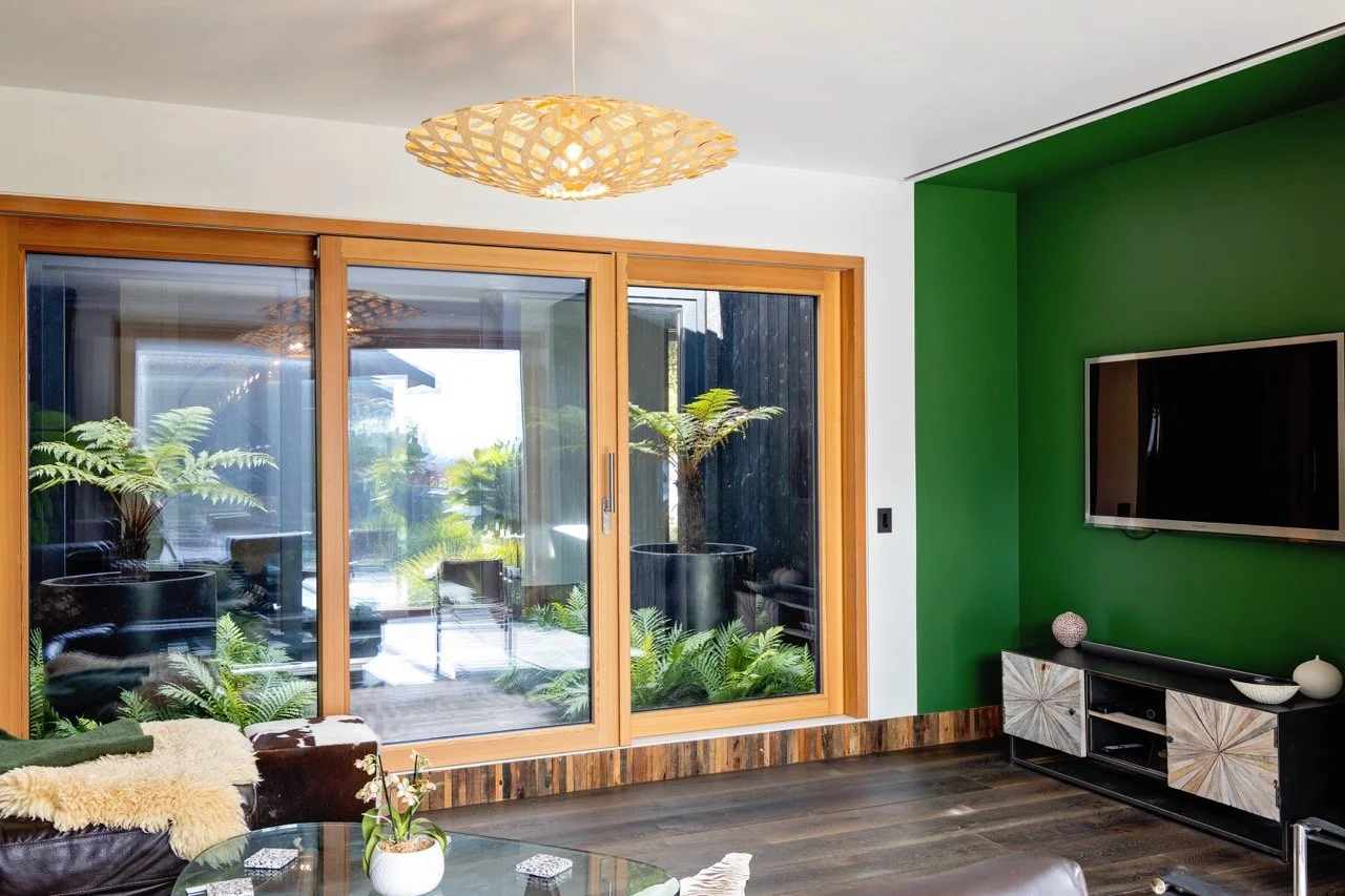 Living room with green accent wall, modern TV stand, flat-screen TV, sliding glass door with wooden frame, plants outside, and patterned light fixture hanging from the ceiling.