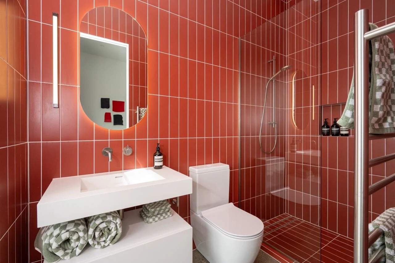 Modern bathroom with red tiled walls, a white sink with a mirror above, a toilet, and a walk-in shower with a glass door and a handheld showerhead. Towel racks hold folded towels and bottles of soap or lotion are on a niche.