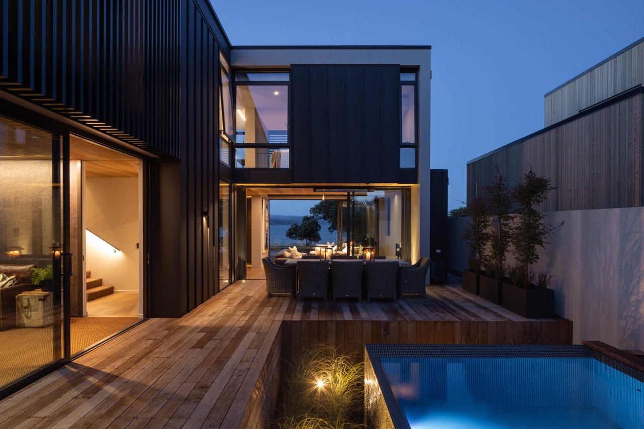 Modern house with outdoor patio, dining area, pool, and large windows during evening.