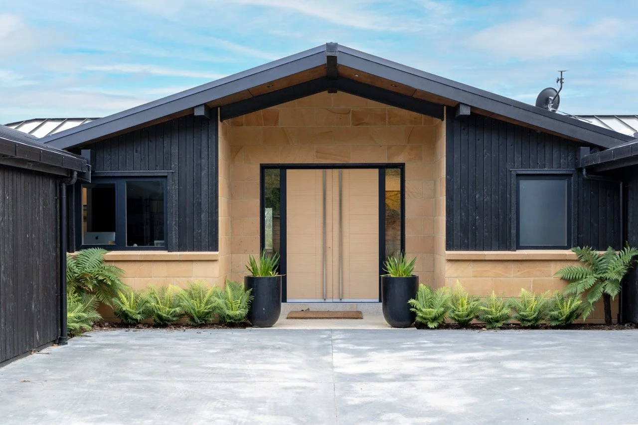 Modern house with black wood siding, tan brick accents, and large black planters with greenery at the entrance, under a blue sky.