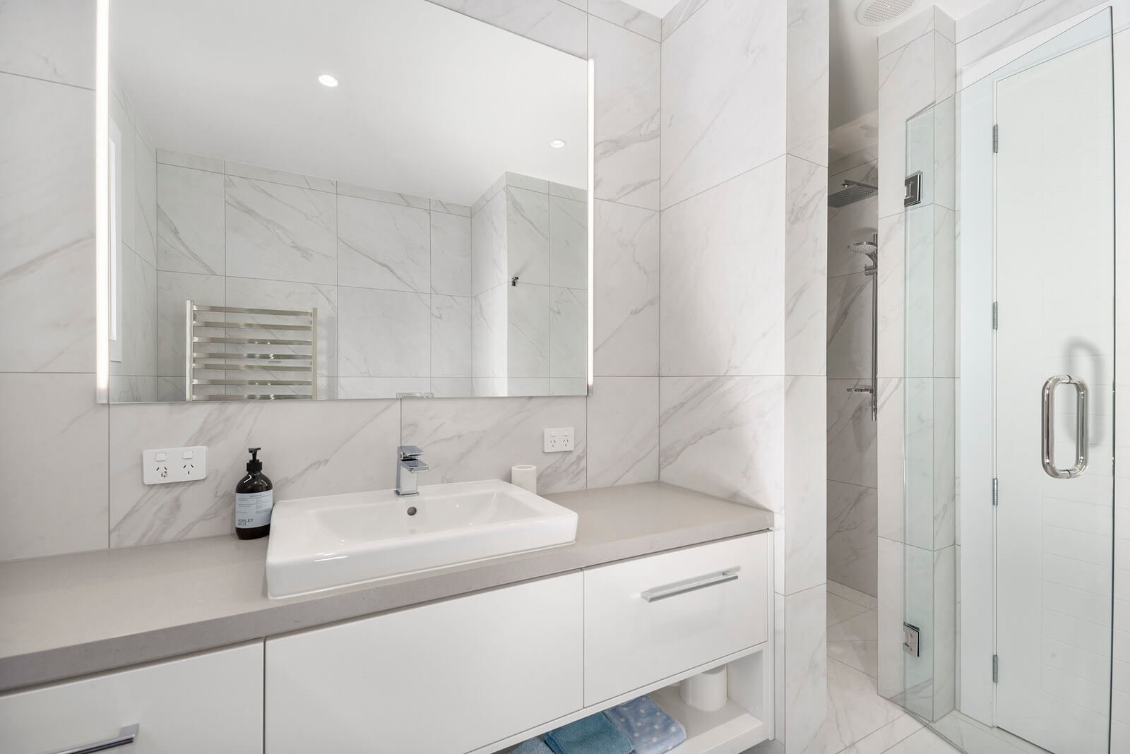 A modern bathroom with white marble walls and a large mirror above a rectangular sink. There is a black soap dispenser and a small bottle on the sink counter. A towel and toilet paper are visible under the counter. To the right is a glass shower encl