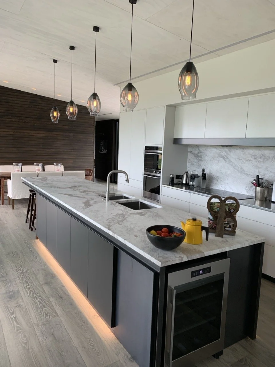 Modern kitchen with a marble island, pendant lights, and white cabinetry. There are bowls of fruit and a yellow teapot on the island. The kitchen has a built-in oven and stainless steel appliances, with wood and marble accents.