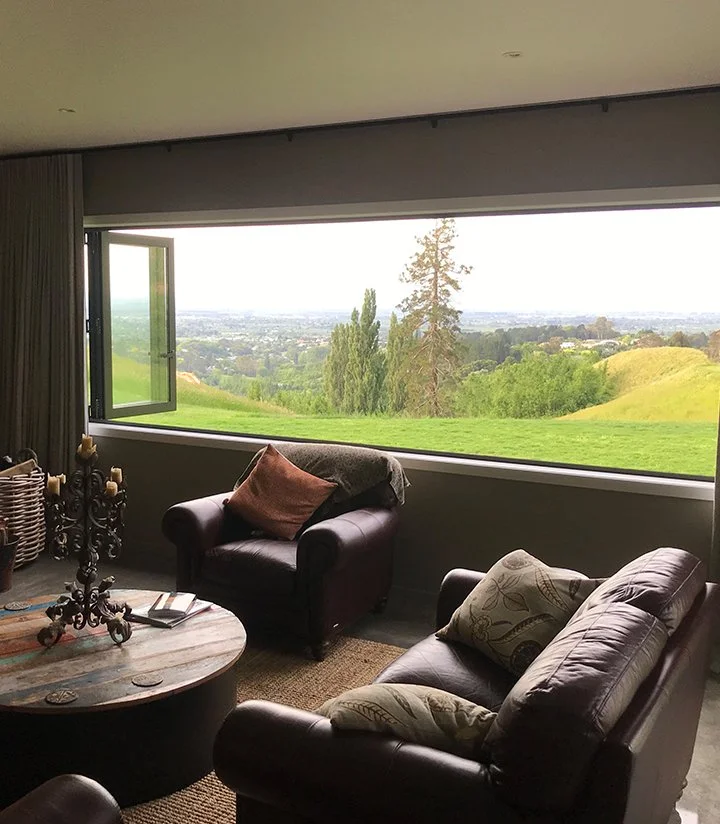 Living room with leather sofa, armchair, coffee table, and large window overlooking green rolling hills and trees.