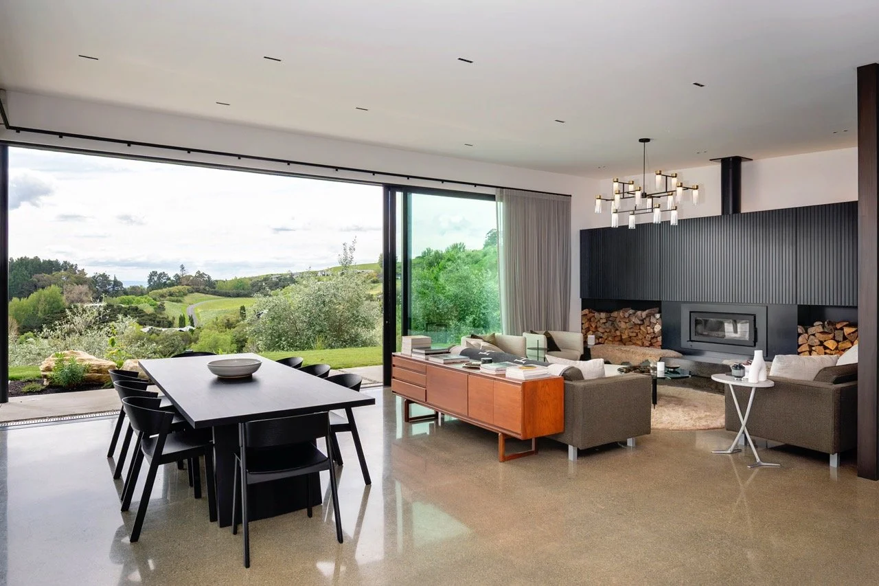 Modern living room with large glass sliding doors showing green hills and trees outside, contemporary furniture, a black fireplace with stacked firewood, and a dining table with black chairs.