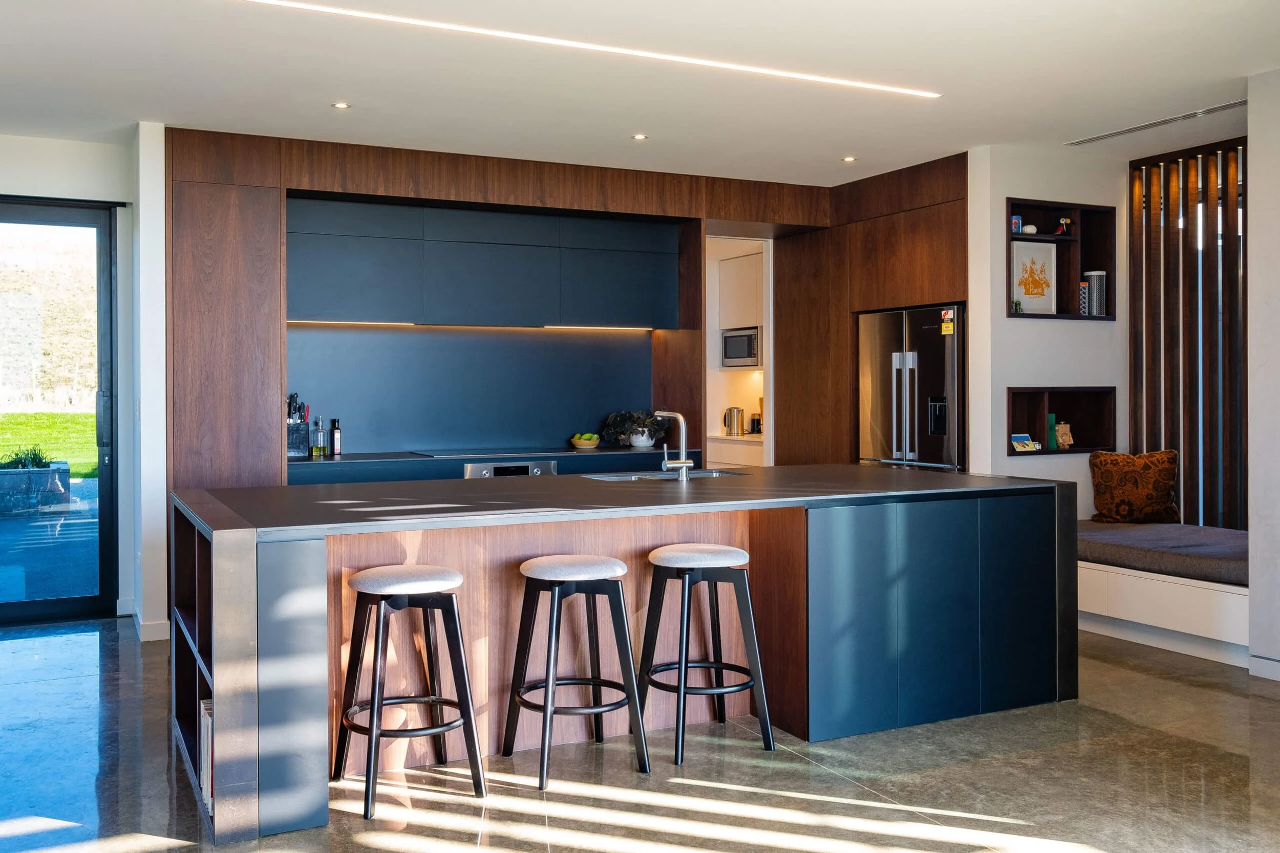 Modern kitchen with dark blue and wood cabinets, a large kitchen island with a wood base and blue sides, three bar stools, stainless steel appliances, and a built-in seating area with cushions.
