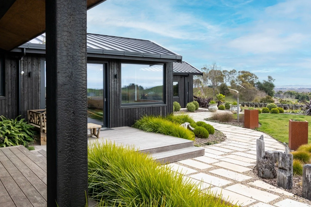 Modern black house with large windows, stone pathway, and landscaped garden with green shrubs and trees in the background.