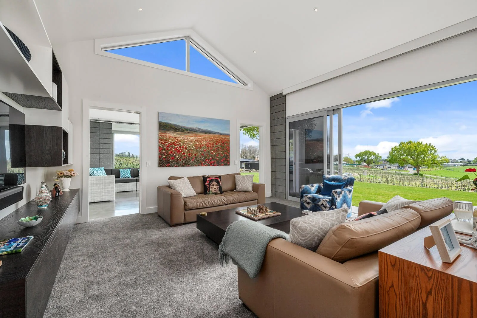 Living room with large glass sliding door showing green countryside and trees outside, modern furniture including brown and beige sofas, a blue armchair, and artwork on the wall, with neutral tones and natural light.