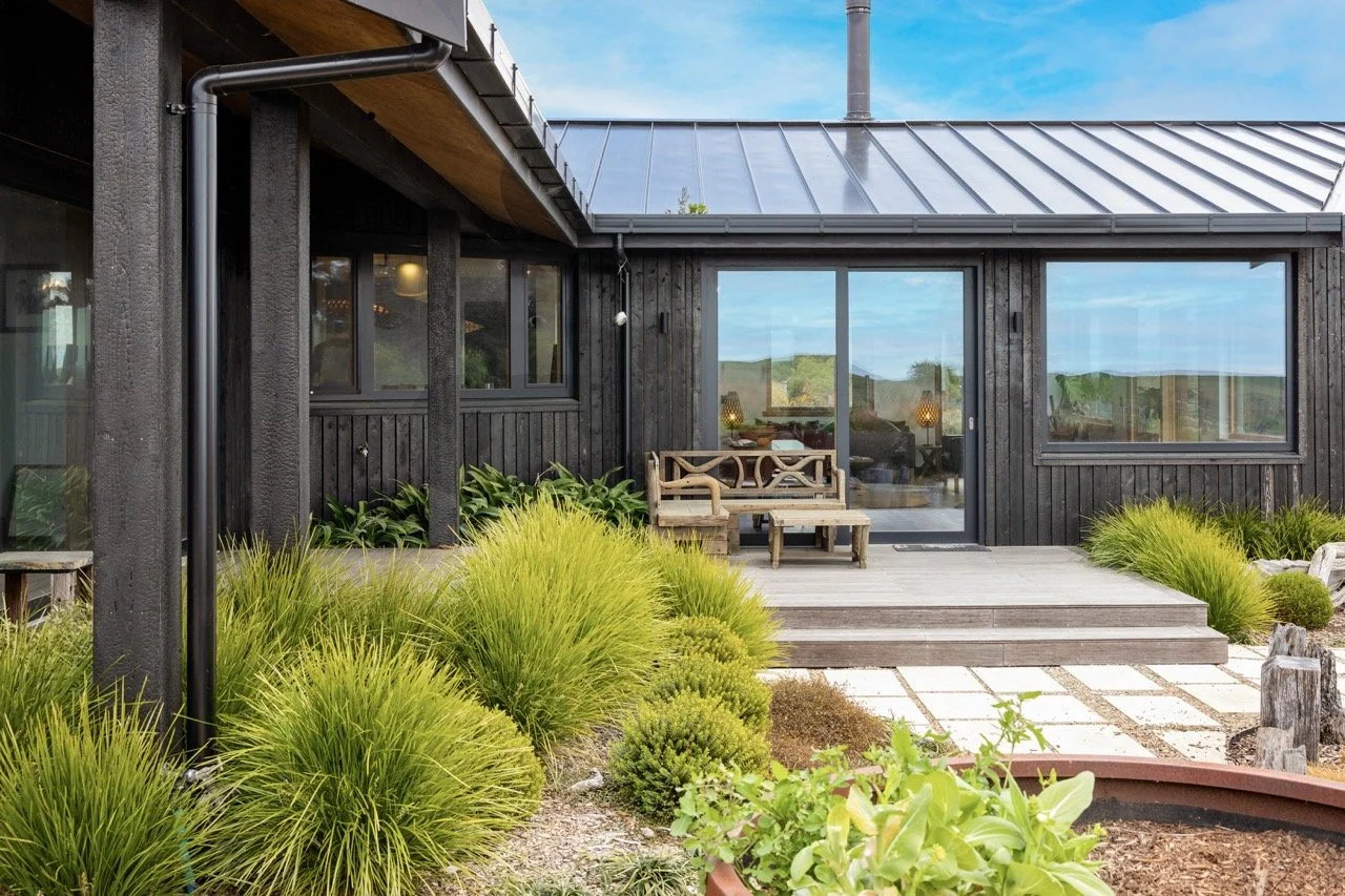 Modern black wooden house with large glass sliding doors, a small wooden porch with seating, surrounded by green plants and landscaping, under a partly cloudy sky.