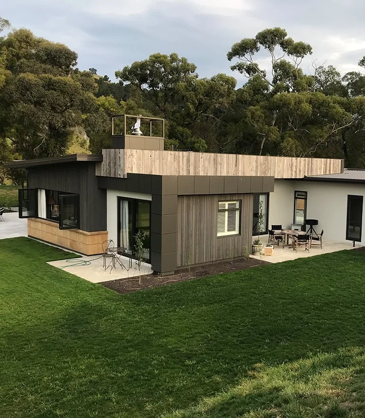Modern house with flat roof, wooden and gray exterior panels, surrounded by green lawn and trees.