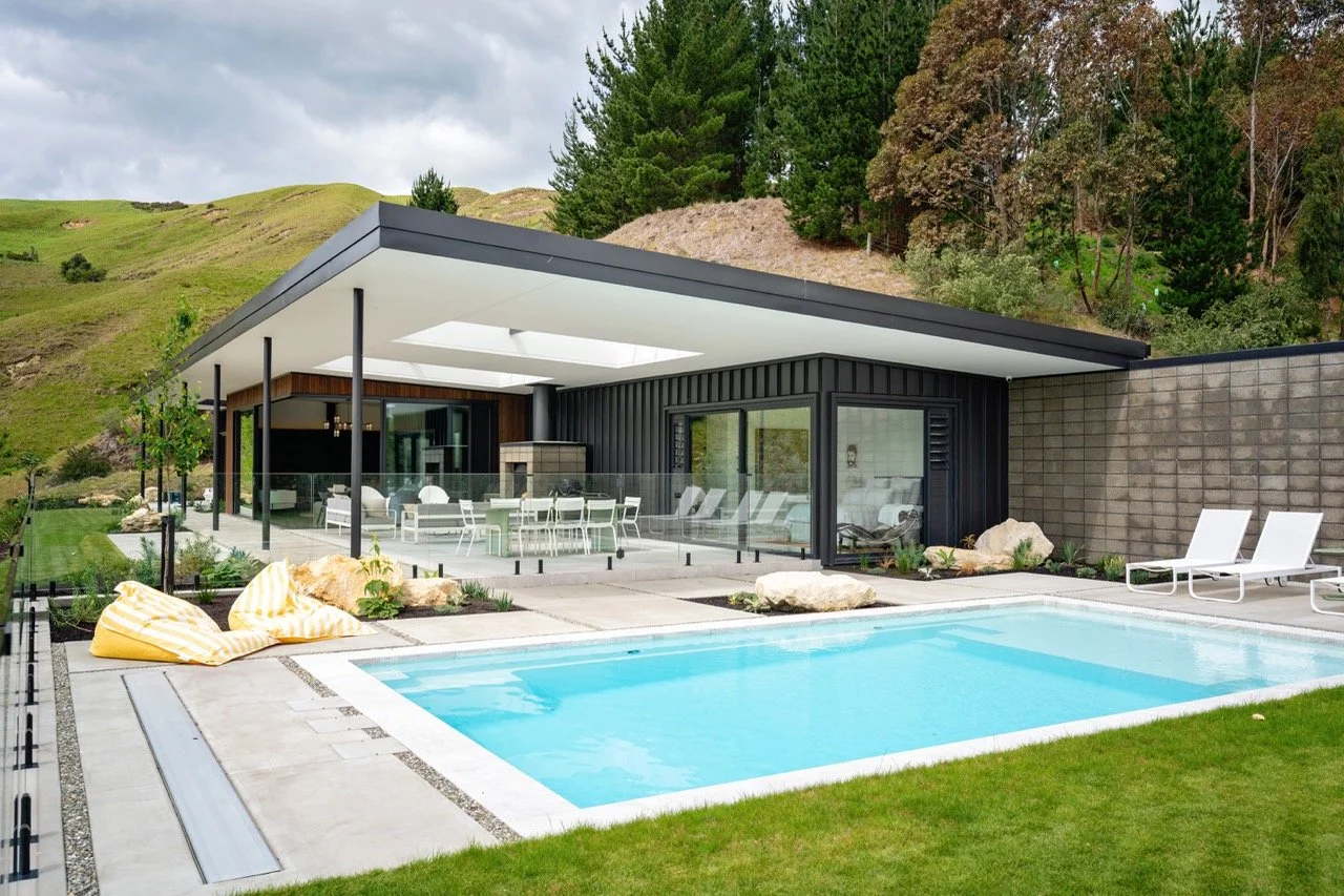Modern house with outdoor swimming pool, lounge chairs, and patio area surrounded by greenery and hills.