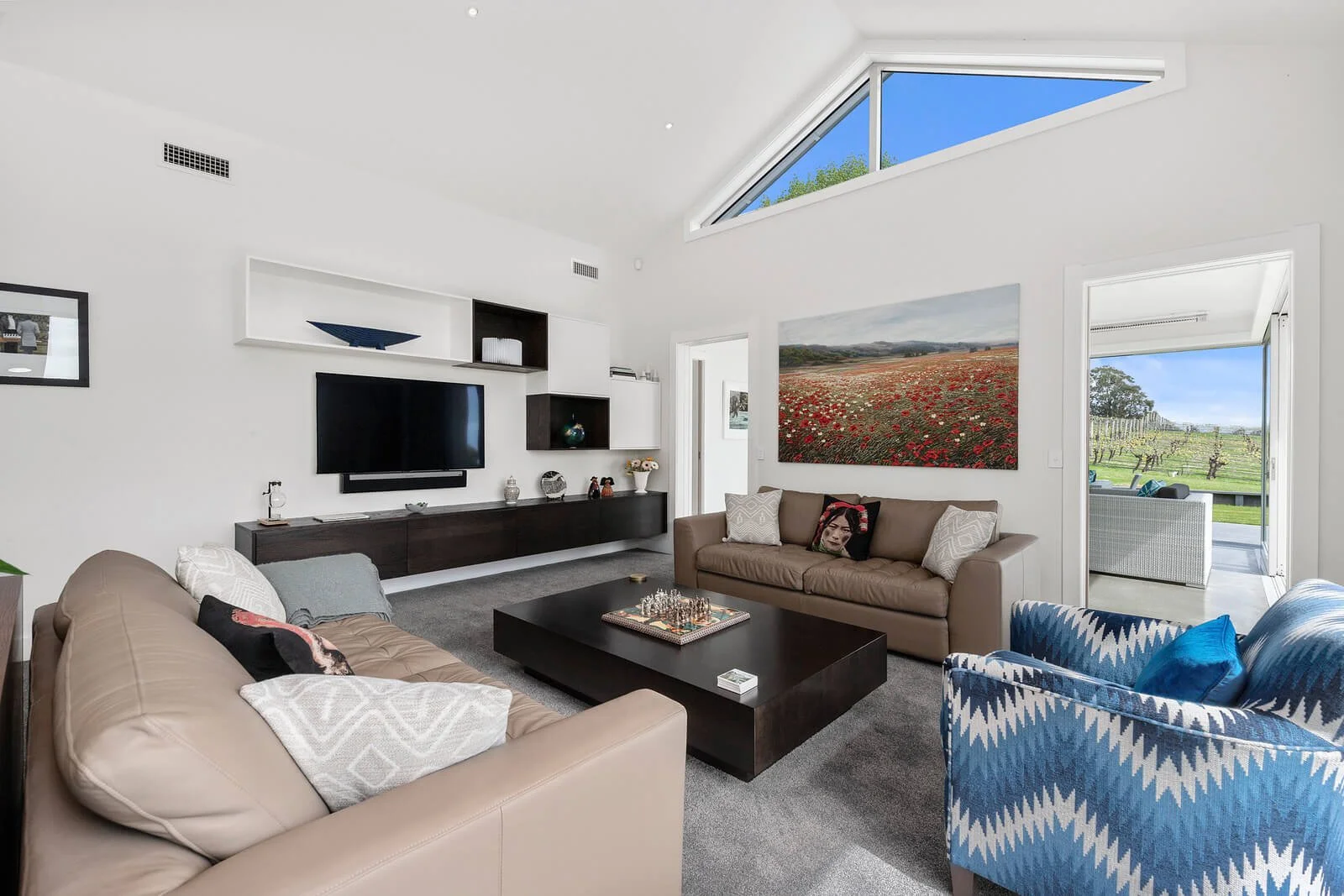 Living room with beige and brown sofas, a black coffee table with a chess set, a wall-mounted TV, and open door leading outside to a patio and a vineyard. Large windowed ceiling shows blue sky and greenery.