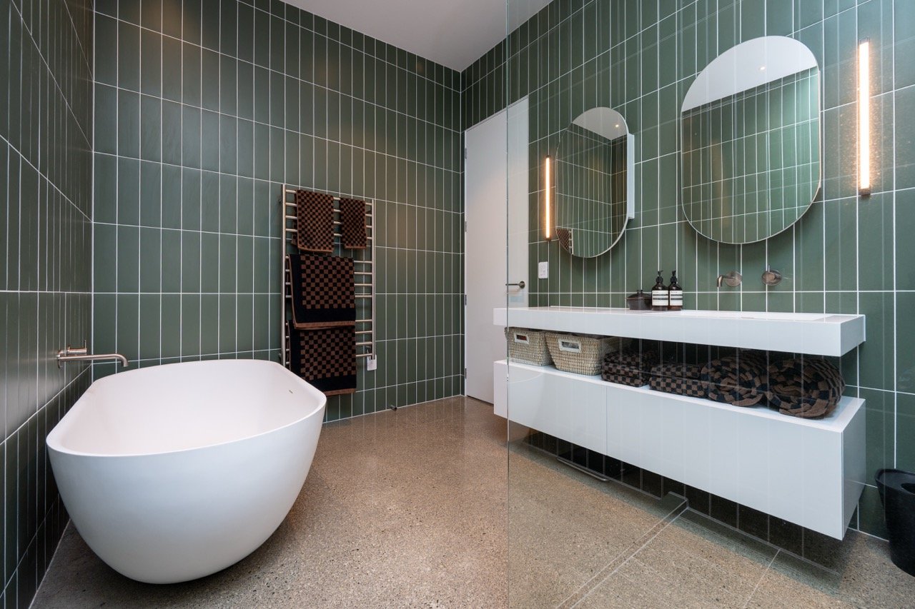 Modern bathroom with green tiled walls, a white freestanding bathtub, a floating white vanity with two oval mirrors, and baskets inside open storage.