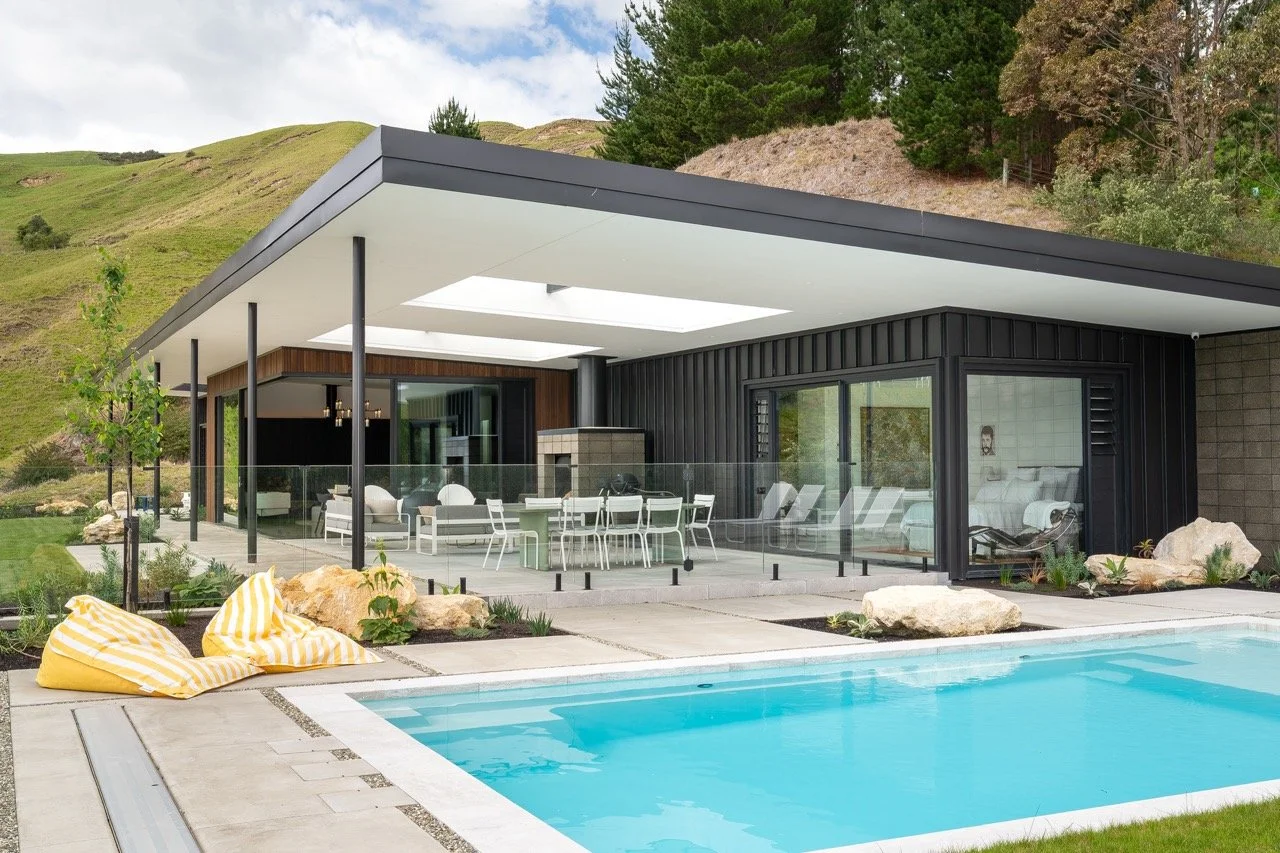 Modern house with outdoor pool, patio furniture, and glass railing, in a natural landscape with green hills and trees.