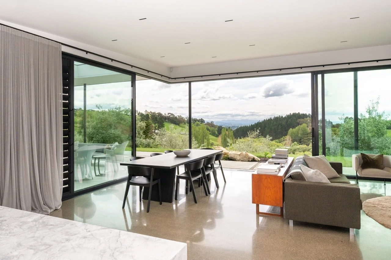 Modern living room with large glass windows overlooking a scenic green landscape with trees and hills, featuring a dining table, chairs, and a sofa.