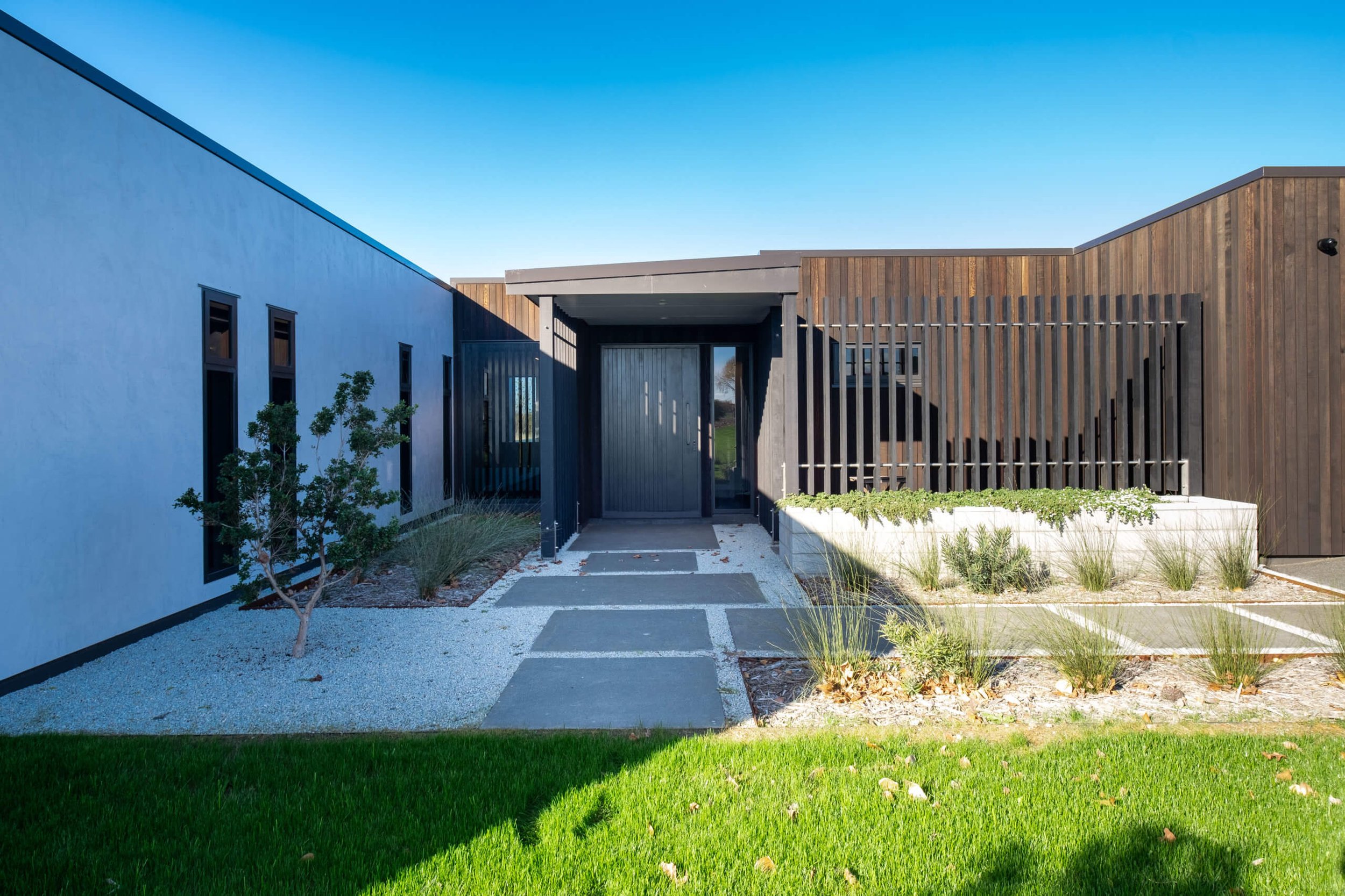Modern house exterior with a pathway leading to the front door, featuring a mix of white and wood paneling, a small tree, and landscaped plants.