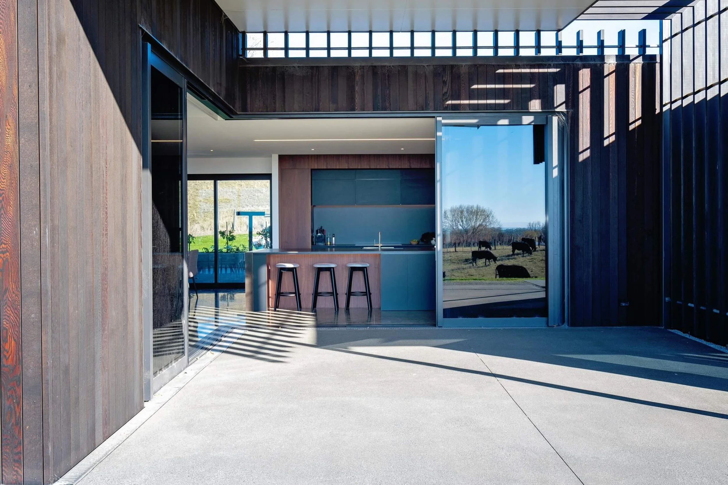 Open-concept modern kitchen with bar stools, dark cabinetry, and large glass window overlooking a pasture with cows.