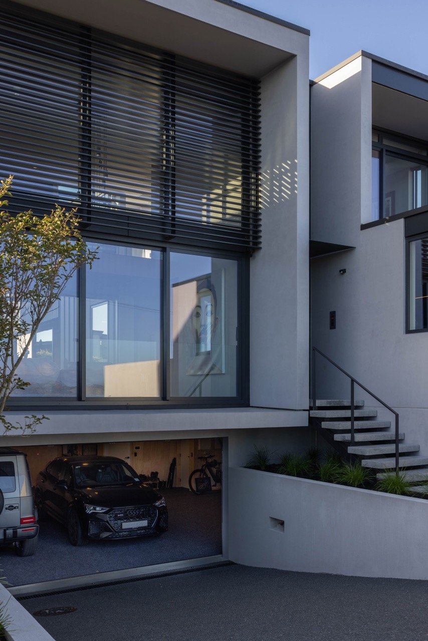 Modern two-story house with large glass windows, black window shutters, and a garage with two cars. Exterior features include concrete stairs, black railings, and minimal landscaping with small plants and a tree.