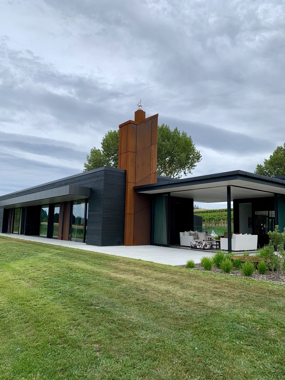 Modern house with black and rust-colored exterior, large glass windows, outdoor seating area with white couches, green lawn, and cloudy sky.