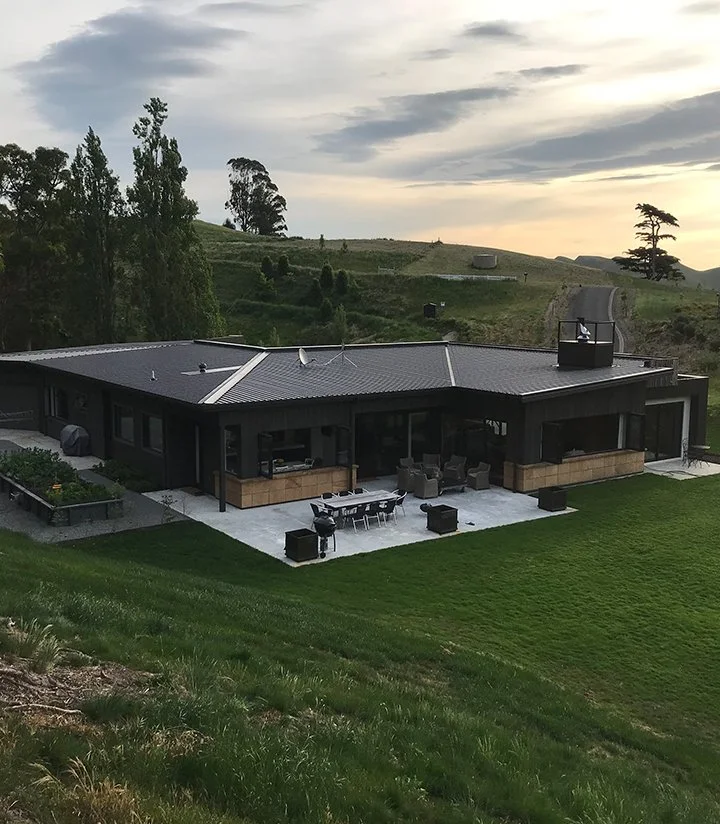 A modern black house with a patio, outdoor seating, and a grill, situated on a grassy slope with rolling hills and trees in the background under a cloudy sky.