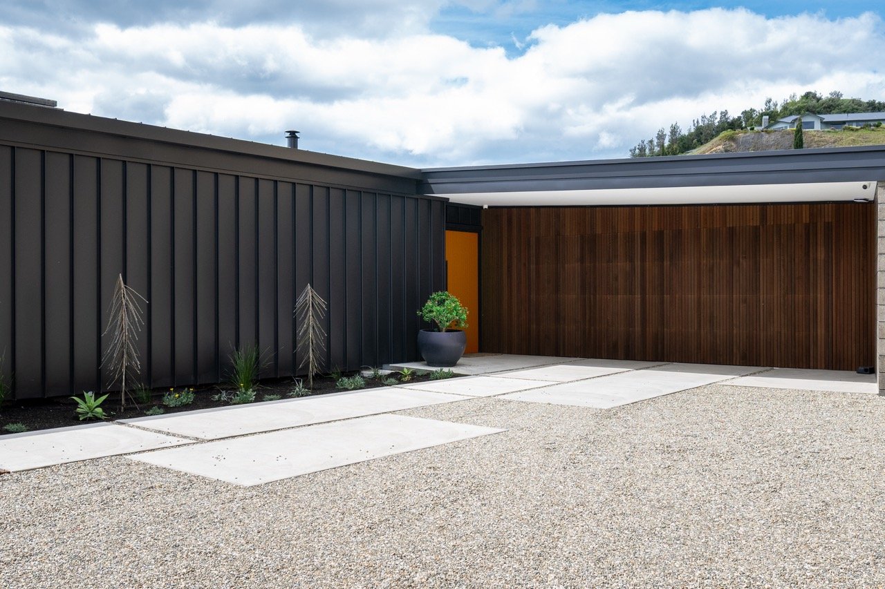 Modern house exterior with black metal siding, wooden garage door, concrete driveway, potted plant, and landscaped garden with small plants under a cloudy sky.