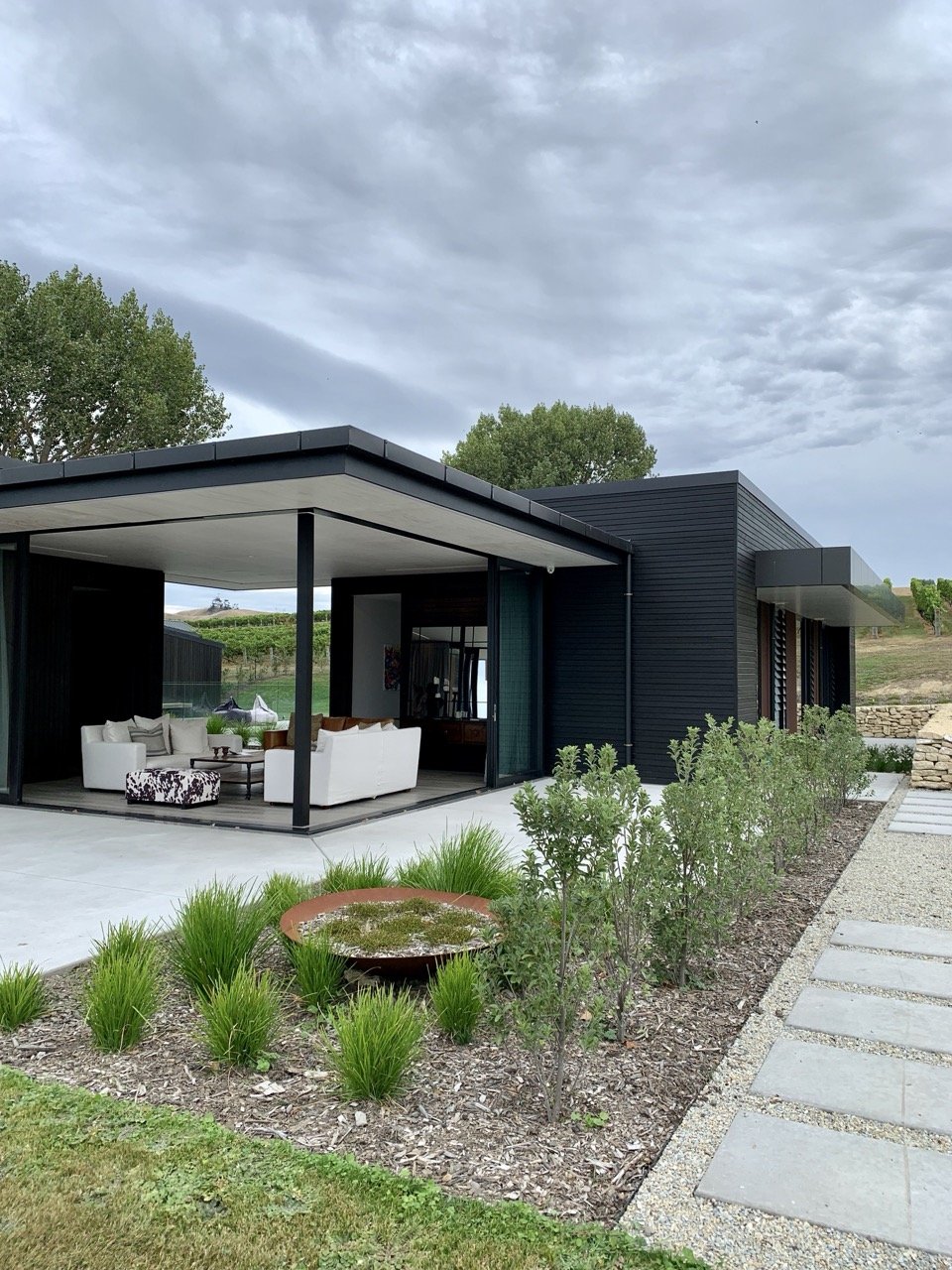 Modern house with black exterior walls, large outdoor seating area with white sofas, lush greenery, gravel pathway, and cloudy sky.
