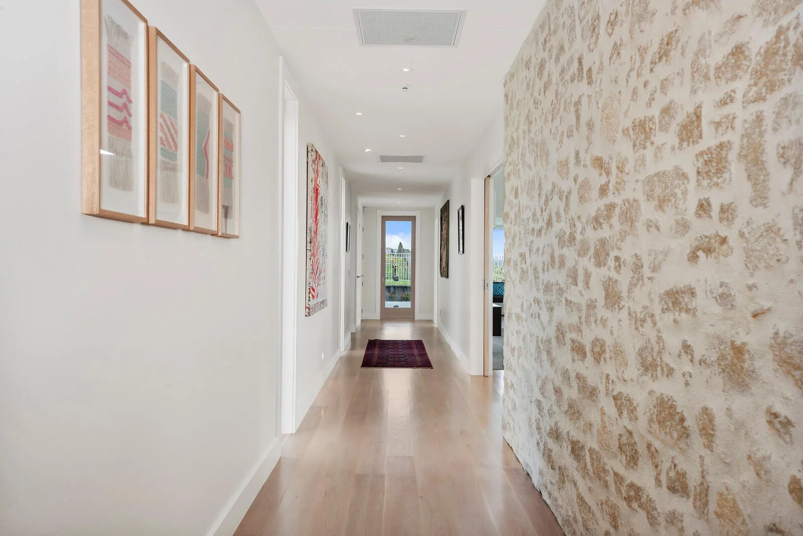 Long bright hallway with artwork on the left wall, a textured wall on the right, and a door at the end with a viewing balcony beyond.