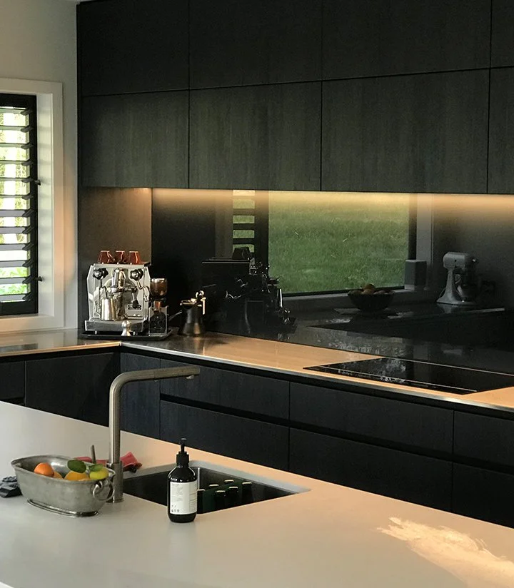 Modern kitchen with dark cabinetry, white countertop, and a window overlooking a lawn. There is a coffee machine and other kitchen appliances on the counter, and a bowl of fruit on the island.