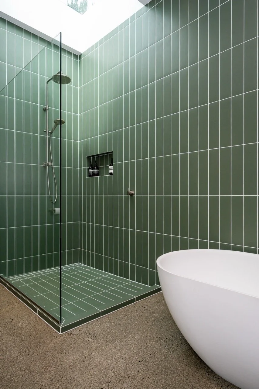 Modern bathroom with green tiled shower area, a built-in shelf with black bottles, a glass shower partition, and a white freestanding bathtub, with a textured brown floor.