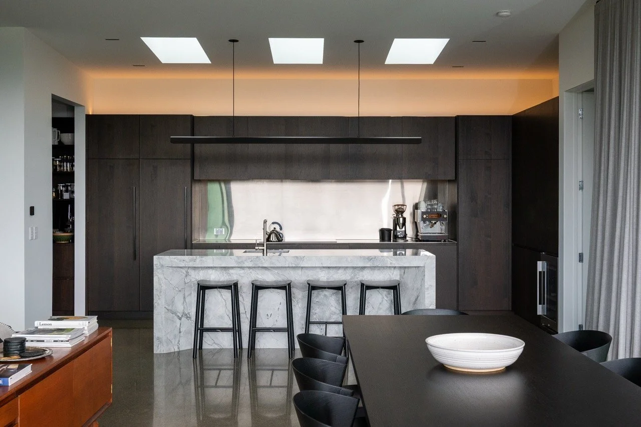 Modern kitchen with dark cabinetry, a marble island, and black bar stools, with a dining table and chairs in the foreground.