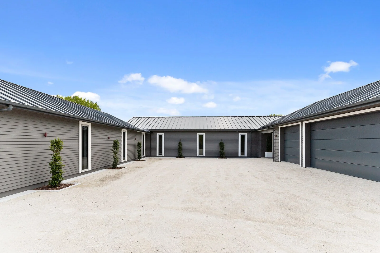 A modern residential courtyard with gravel ground, gray siding buildings, narrow windows, and a gray metal roof, under a blue sky with a few clouds.