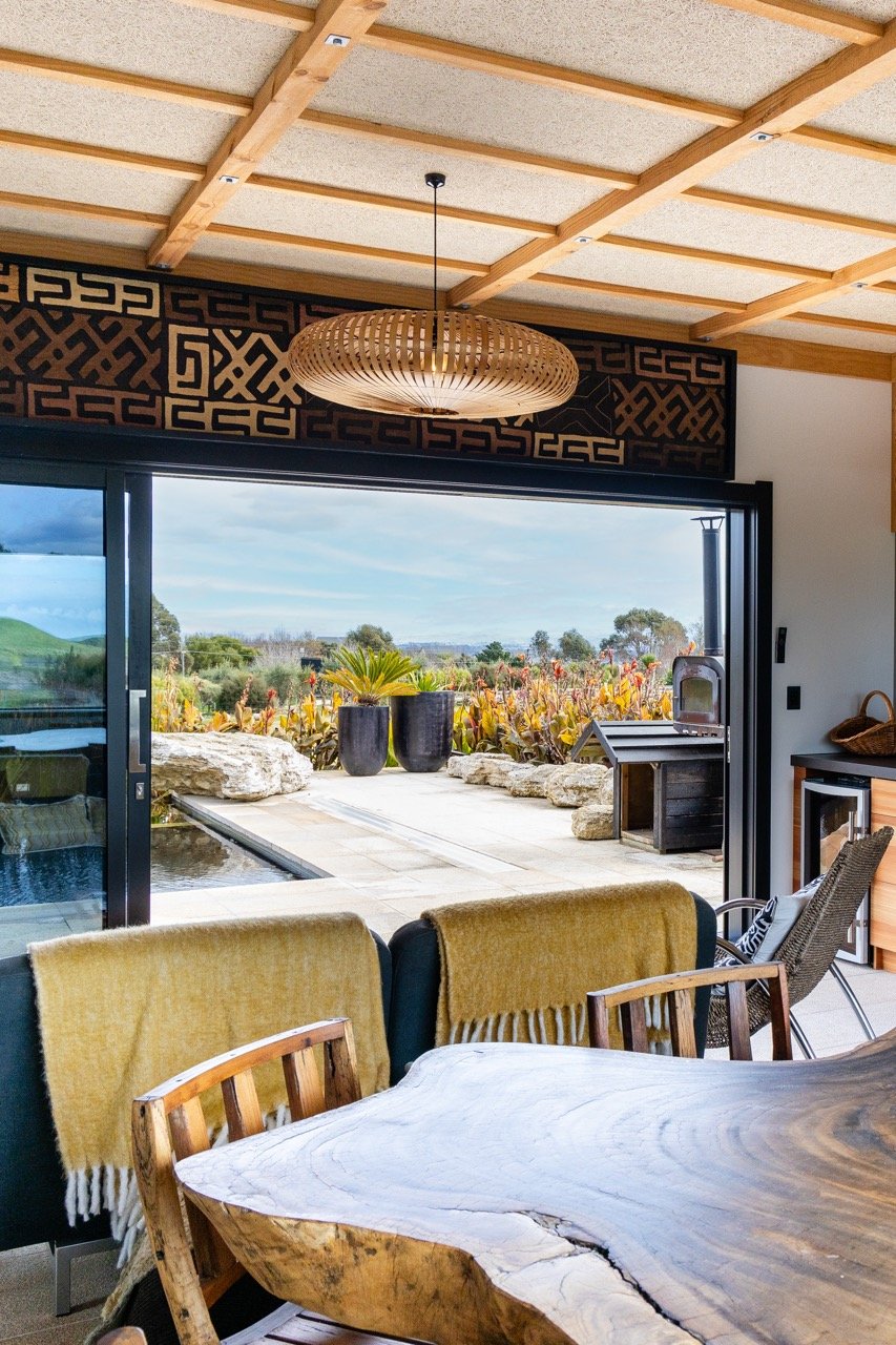 Inside a modern house looking out to a patio and garden, with a wooden dining table, chairs, and sliding glass doors.