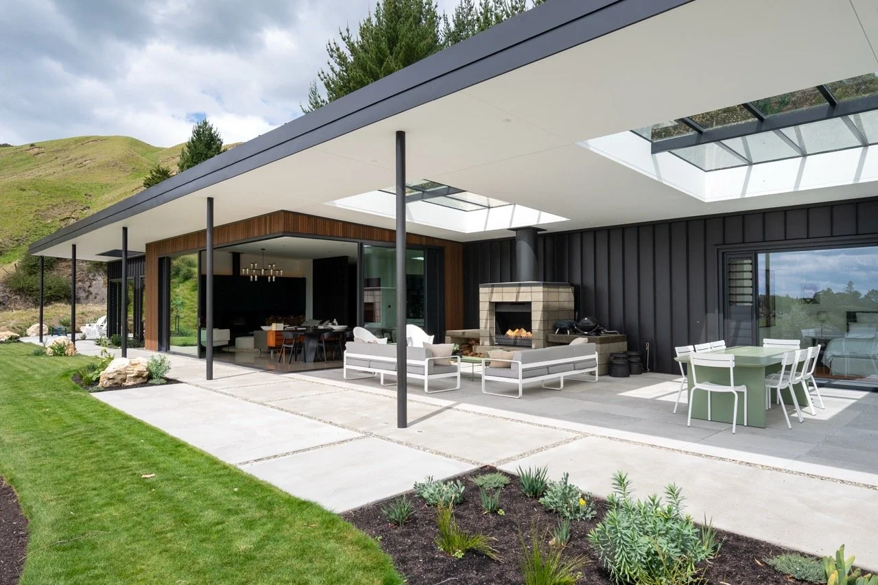 Modern house with large outdoor patio, outdoor fireplace, dining table with chairs, and seating area with white furniture, located in a green landscape.