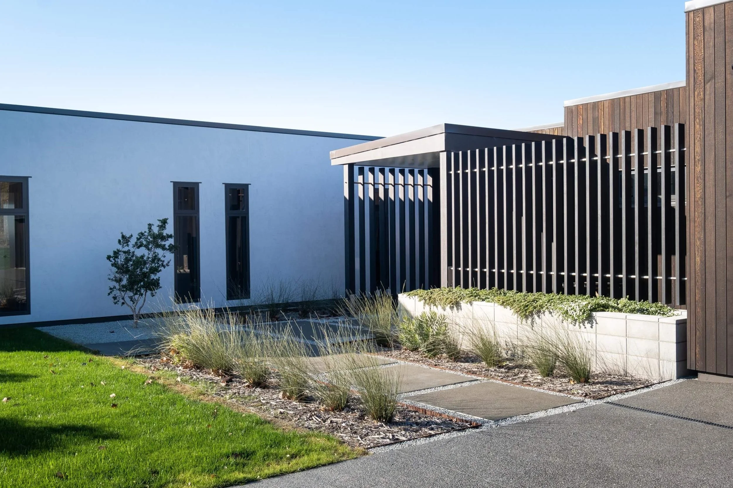 Modern building exterior with a white wall, tall narrow windows, landscaped garden, and wooden slatted structure, under a clear blue sky.