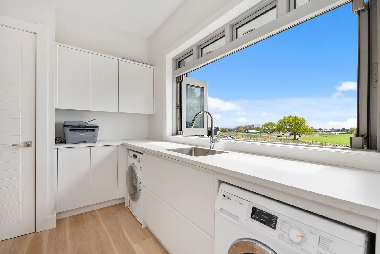 Bright laundry room with white cabinets, a window with a view of a sunny countryside landscape, washing machine, and laundry dryer.