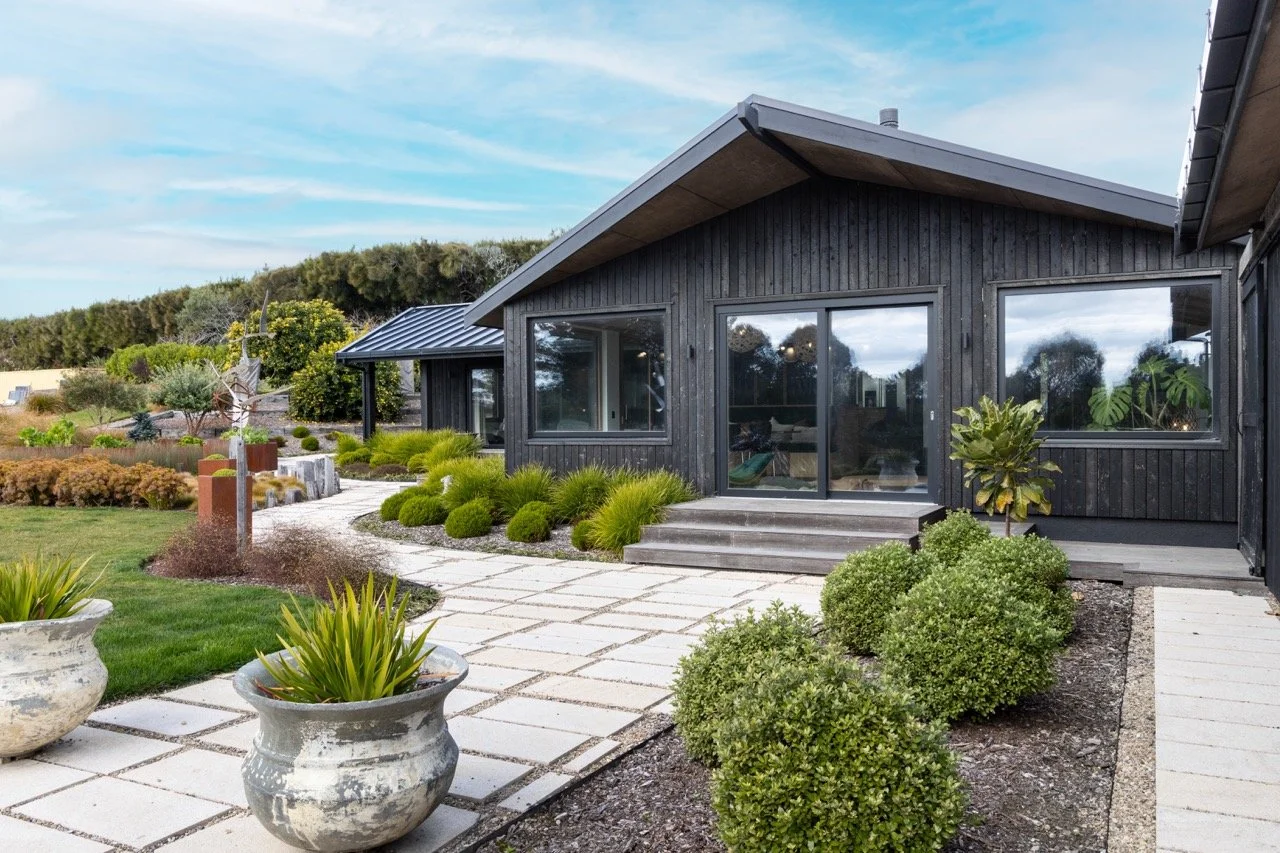 A modern black house with large windows, surrounded by a landscaped yard with potted plants, bushes, and paving stones, under a partly cloudy sky.