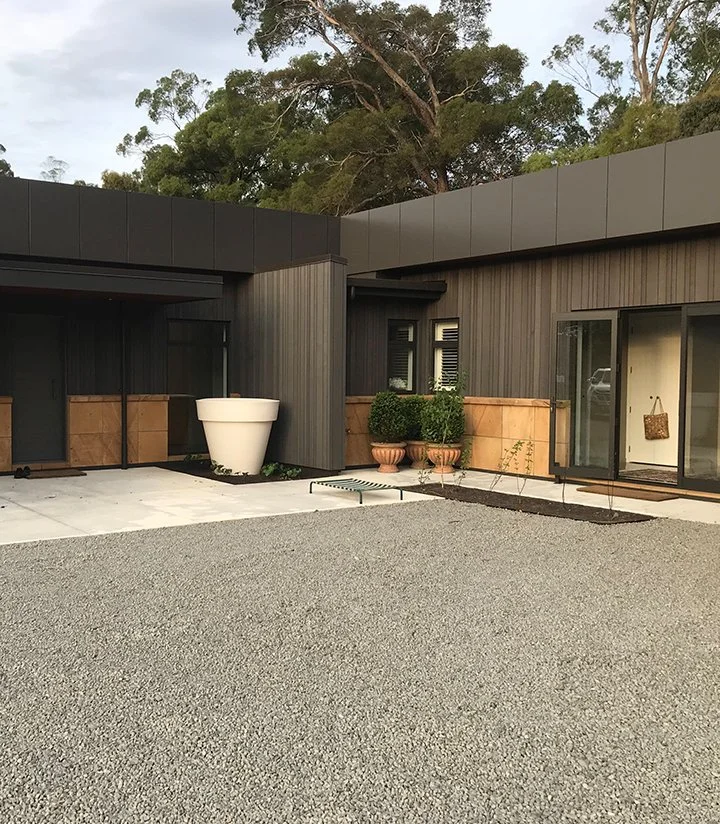 Modern house with dark exterior walls, large white planter, potted plants, sliding glass door, and gravel backyard, surrounded by trees.
