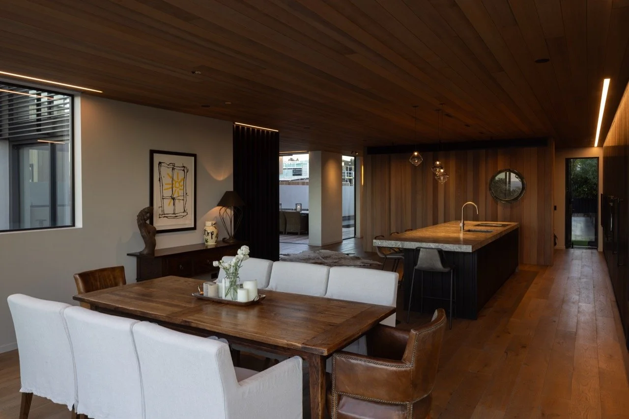 Modern open-concept living and dining area with wooden accents, a dining table with white chairs, a kitchen island with pendant lights, and large windows allowing natural light.