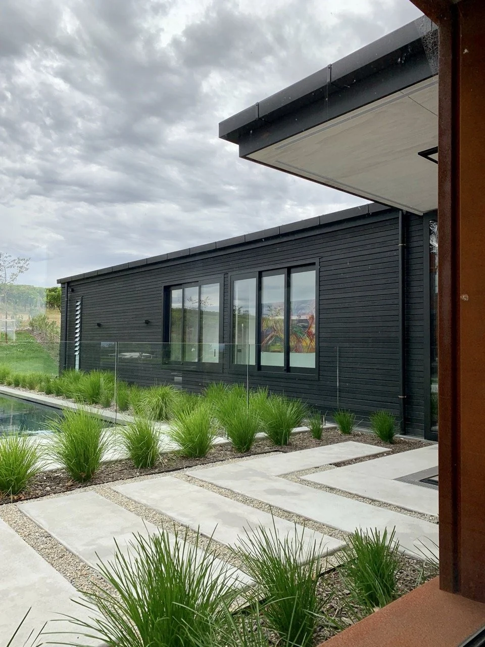 Modern house exterior with black siding, large windows, concrete pathway with grass and plants, cloudy sky.
